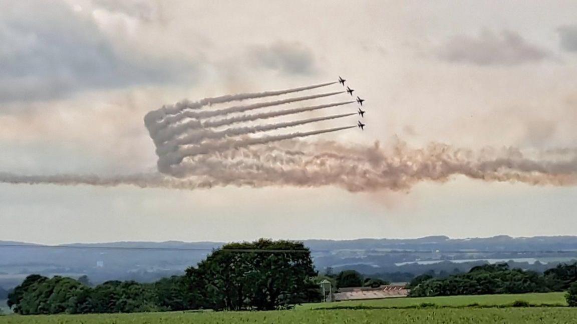 Five planes bank in the sky, trailing long plumes of smoke behind them, over green trees and fields.