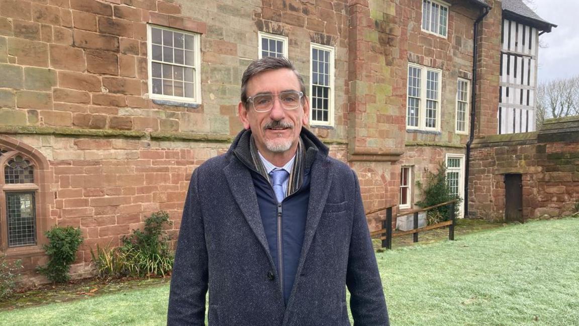 A man with brown hair and a grey beard with glasses and a blue coat standing on a grass lawn in front of an old red brick building with white-framed windows