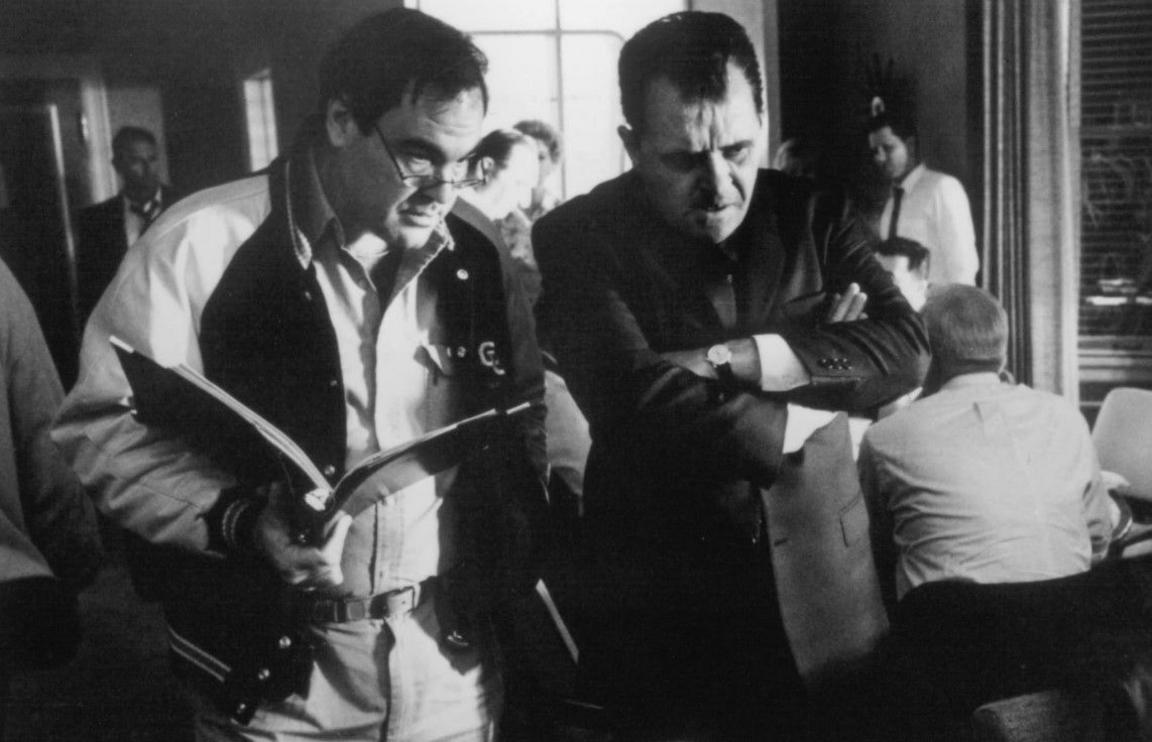 Behind the scenes black and white photo showing:
The director, Oliver Stone (L) with glasses and holding a file  talking to Hopkins in character in a suit with his arms closed

