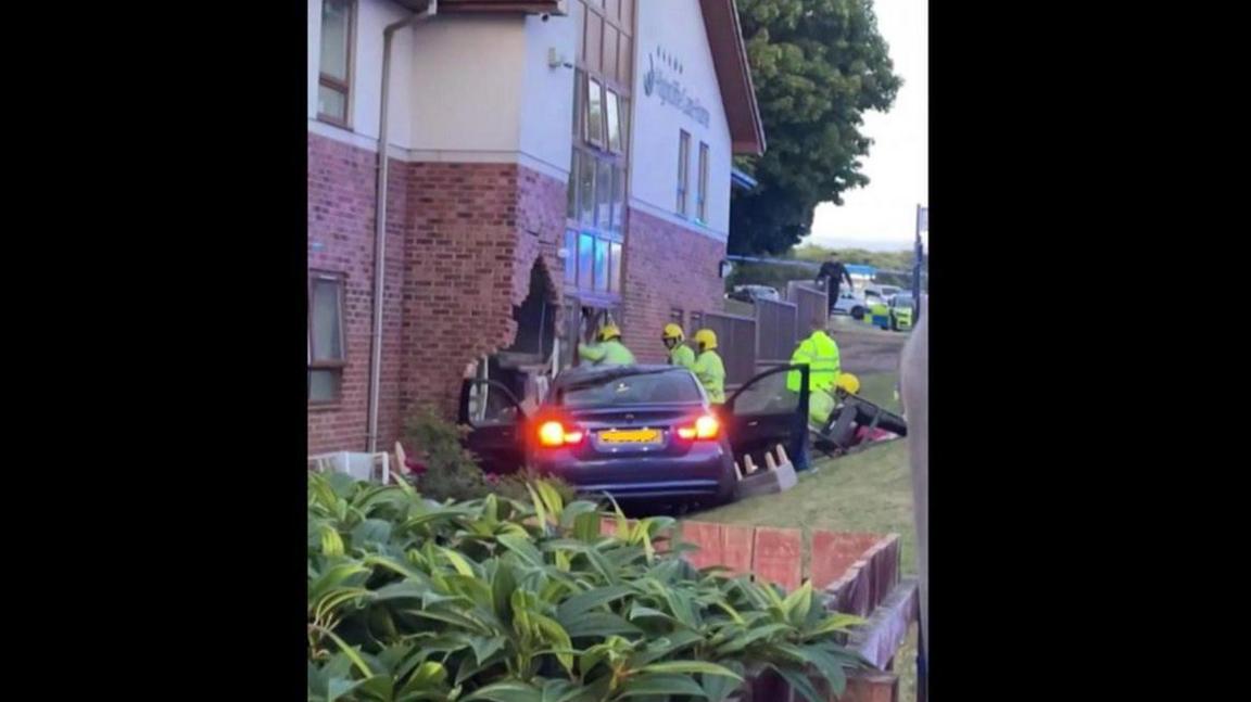 A blue BMW car with its doors open and its rear headlights on, halfway through a brick building. Firemen in hi-vis jackets surround the car.