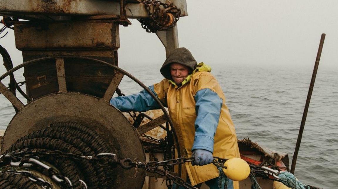 A fishermen pulling a large metal chain while on a boat out at sea. His face is tense from the effort. He is wearing yellow and blue water-proof clothing with a hood over his head. The sea behind him is grey.