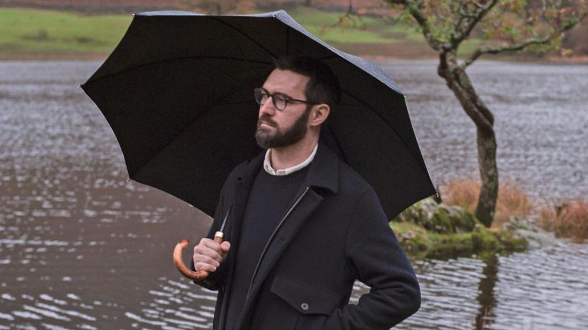 Lee Paton. He has dark hair and a beard and is wearing a black coat and jumper. He is holding a black umbrella above his head and is standing in front of a lake.
