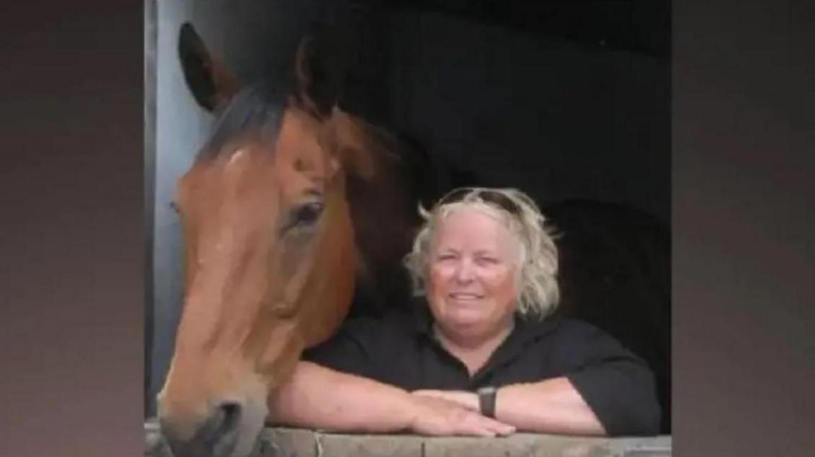 Wendy Buckney looking out from a stable door alongside a horse. She has blonde hair and wears a black top and her arms are folded, resting on the stable door