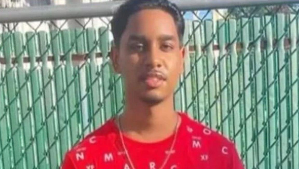 Shawn Seesahai wearing a red t-shirt and neck chain. He is standing in front of a green fence.