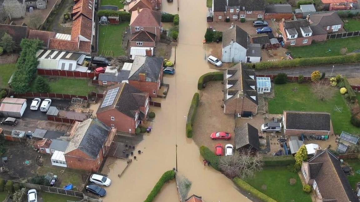 An aerial view of a residential street with rows of homes on either side inundated with brown flood water