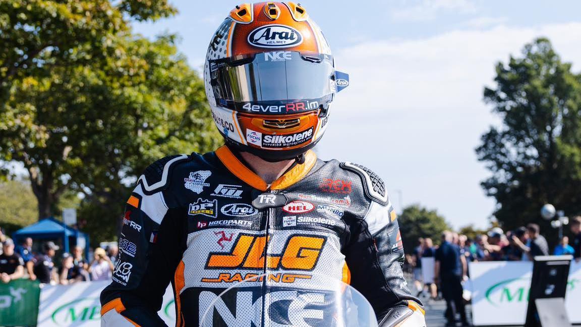 Jamie Williams sitting on his bike on the start line. He is wearing black, white and orange leathers and a matching helmet. He has JLG Racing written across the front of his leathers.