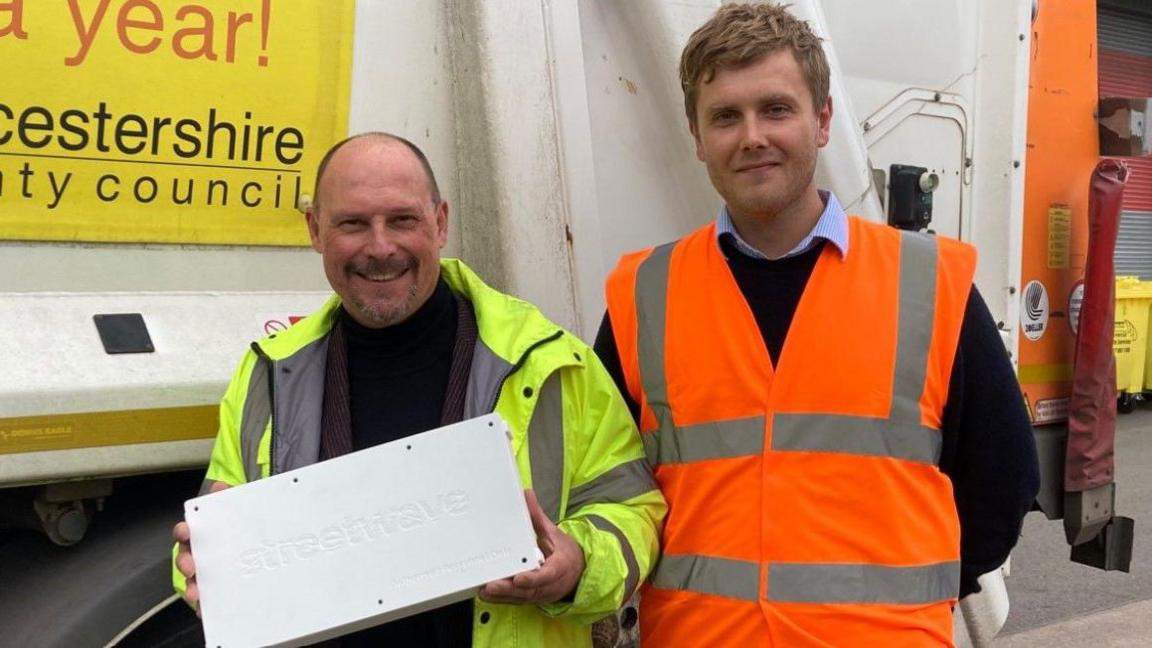 Two men stand in front of a bin lorry. On the left is a smiling man in a yellow hi-viz jacket holding a white box that has the word streetwave embossed in it, and on the right is a man wearing an orange hi-viz vest