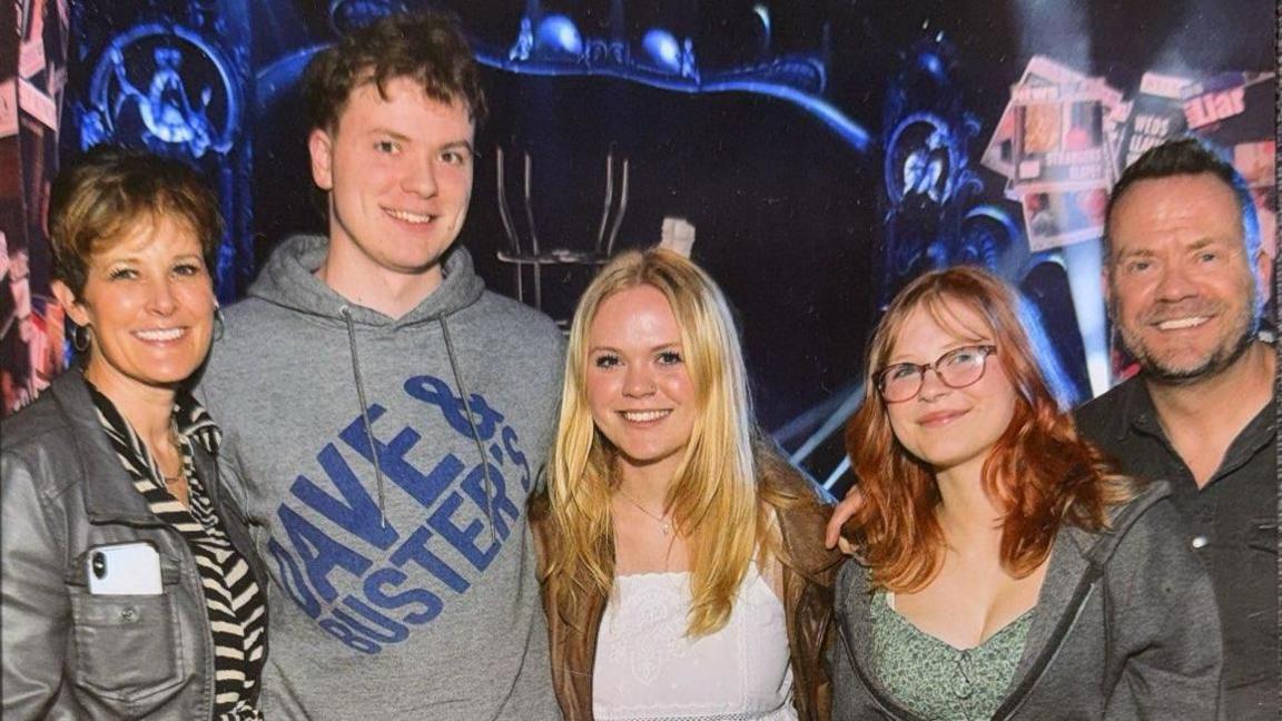 The Waters family pose for a photo in a line. They have their arms around each other and are smiling at the camera.