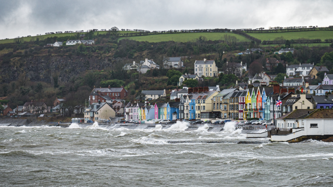 A stormy sea batters a prominate along which is a row of colourful houses. In the background some green fields and above a cloudy sky