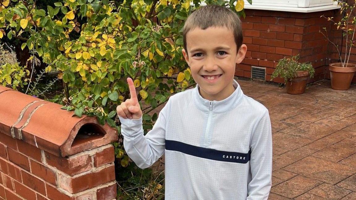 A child is standing on a paved driveway next to a short red-brick wall with a curved top. Behind the wall is a green shrub with yellowing leaves, and in the background, there is a red-brick house with white-framed windows and potted plants near the wall. The child is wearing a light grey long-sleeved top with a dark horizontal stripe across the chest, black shorts, and yellow-and-black trainers. One hand is raised with the index finger pointing upward.