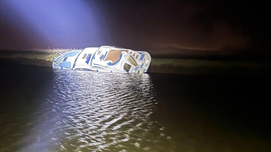 A white boat in a dark river, listing to one side into the water as a bright spotlight shines on it from above.