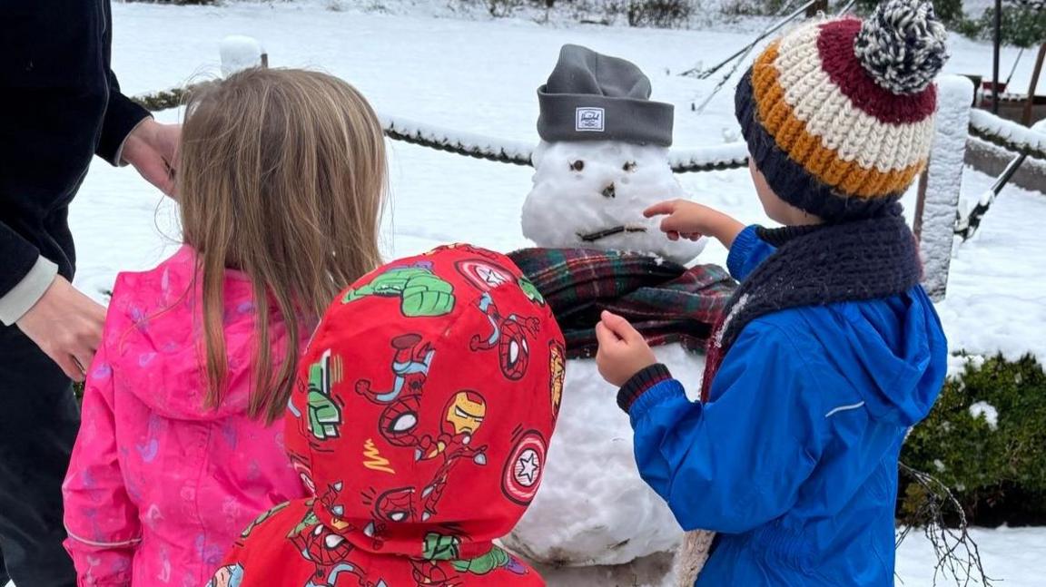 Girl in pink snow suit, boy in superhero snowsuit and boy in blue snowsuit with wooly hat on admire their snowman's face