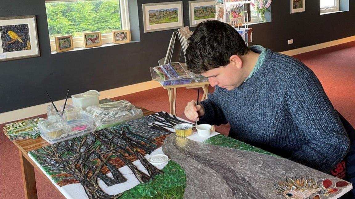 James Owen Thomas creating his piece of art - sitting down painting, after litter was chopped into small pieces and added to the canvas. Paintings are on the walls and by the window.