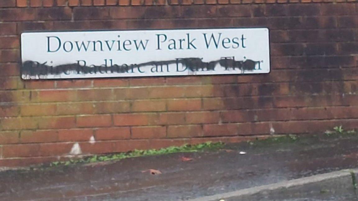 Paint daubed on a black and white street dual-language street sign in north Belfast. It is attached to a red brick wall. 