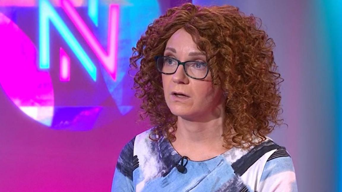 A woman with red curly hair, black-framed glasses and a blue patterned top in front of a pink and blue background