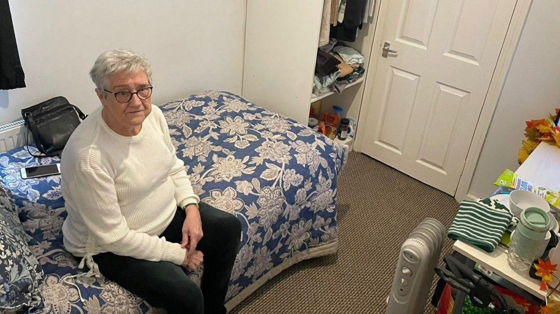 Susan Curtis - a woman in her 70s with short grey hair - sitting on a bed in a small room 