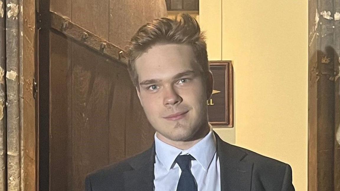 Illia Mitiushnikov, who has fair hair and is wearing a dark suit, standing in the front doorway of an old stone building.  