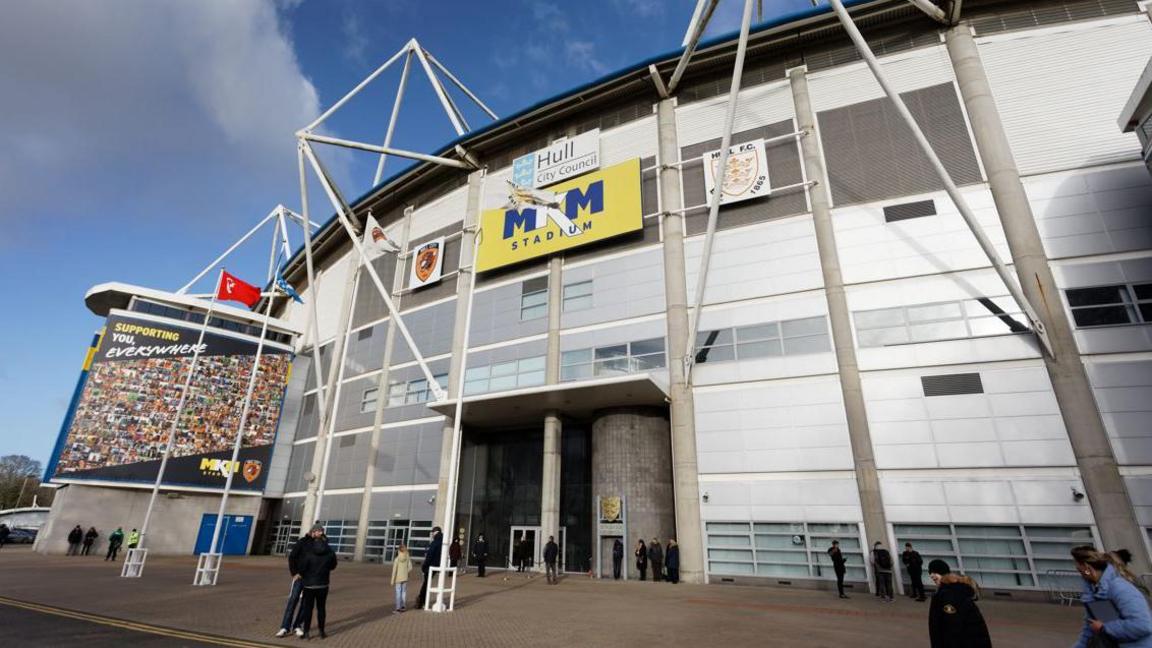 MKM Stadium - a large grey-panelled structure, featuring girders coming out. People are walking past. 