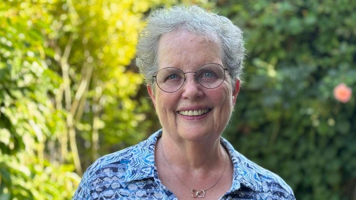 A woman with grey hair and glasses is smiling at the camera. She has a blue top and bushes are blurred in the background.