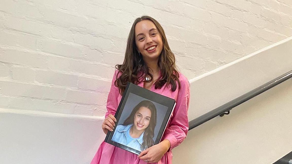 A woman, Chloe, stands on a flight of stairs holding a folder with a picture of her face on it. She is wearing a pink dress and smiling. 