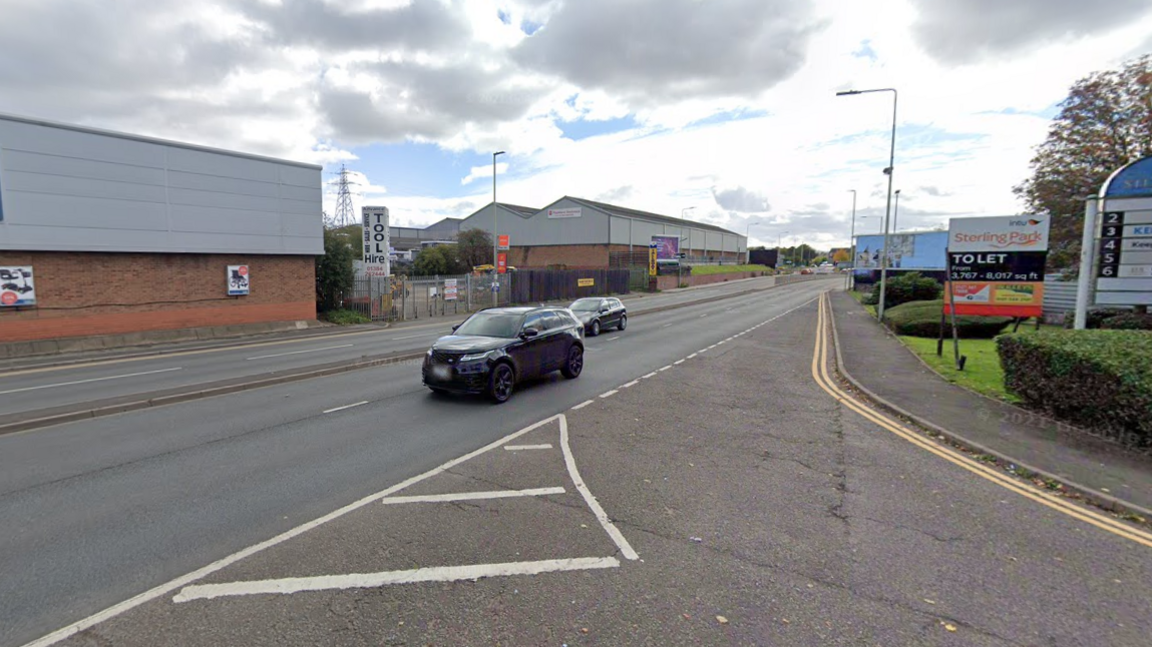 A road with industrial units on either side. Two cars are travelling past and a junction leads off to the bottom right of the image.