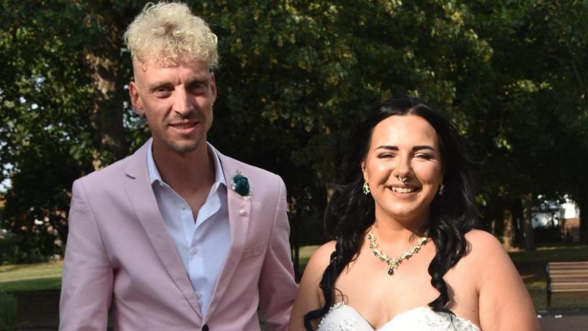 Sammi Hill and her husband Adam Hill. Sammi has black hair which is partially tied back. She is wearing a strapless wedding dress with a gold and green necklace. Adam is standing on the left, wearing a baby pink suit. He is taller than Sammi and has blonde curly hair with short sides. They are standing in front of a large tree.