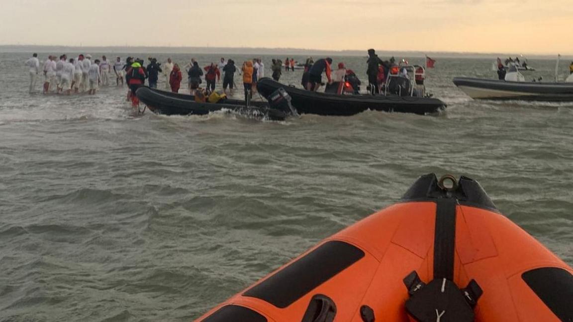 A photo taken from an orange RNLI RIB of cricketers in whites and spectators standing ankle-deep in the sea. The small sandbar on which they stand is surrounded by boats.