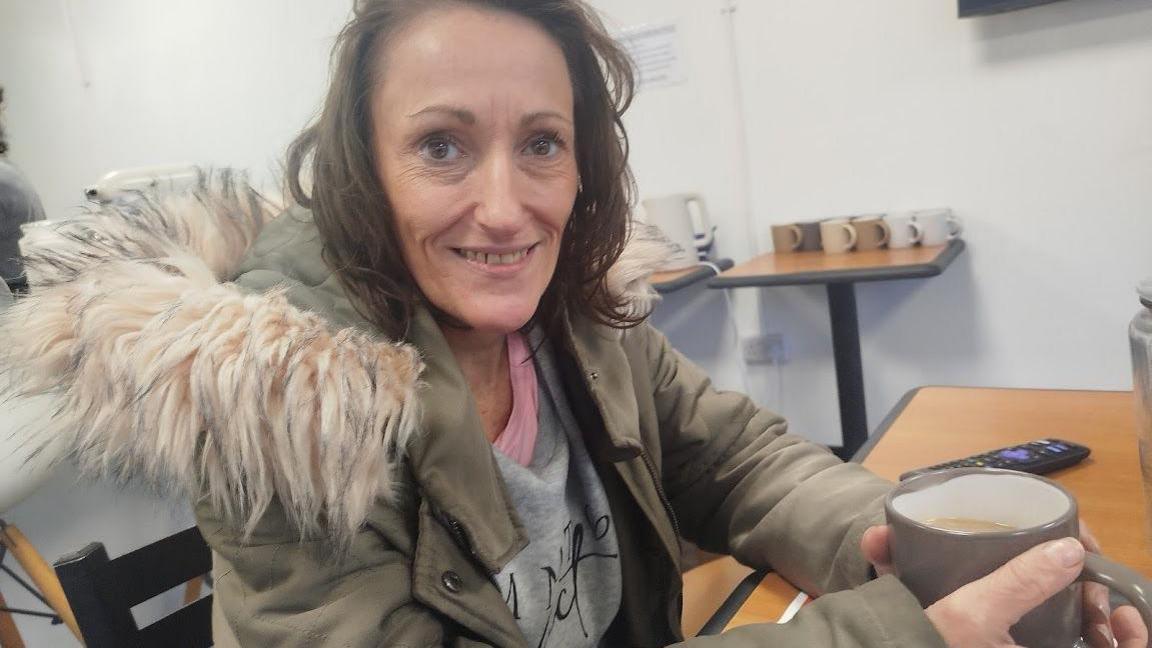 Sarah Falconer has dark brown hair and is wearing a khaki parka. She has her hands cupped round a large mug of coffee.