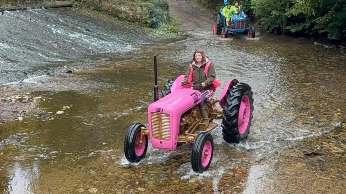A woman drives a pink tractor through a stream.
