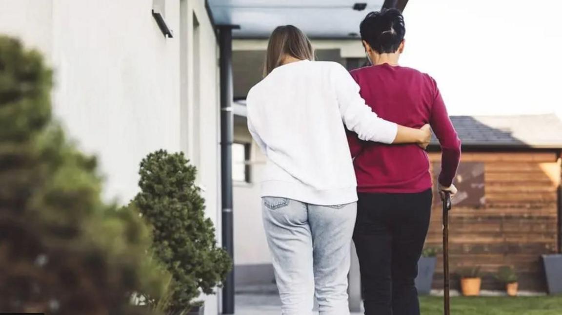 A younger woman dressed in white from behind with her arm around an elder woman in a red top and black trousers using a walking stick. The younger woman seems to be helping the older woman up a pathway to a house.