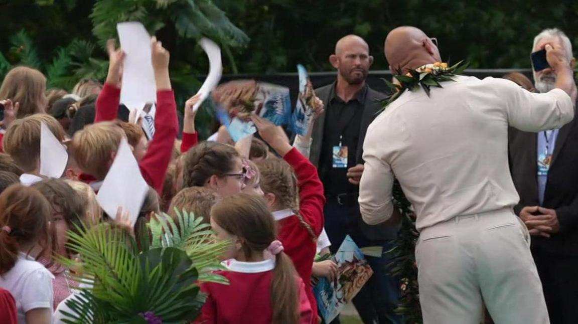 A group of schoolchildren wearing red uniforms standing in front of The Rock – a man wearing a cream jumper and white trousers and sunglasses.