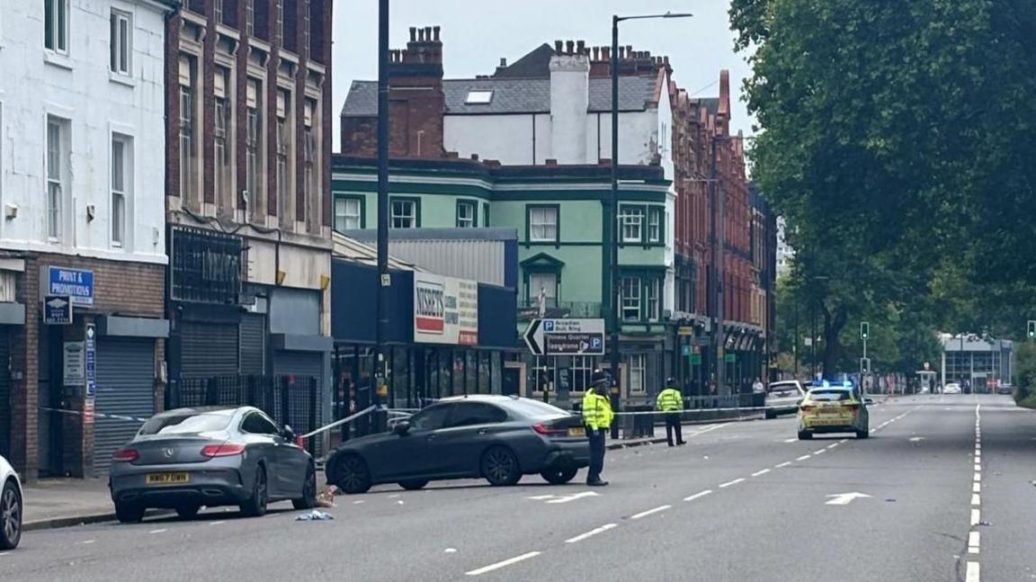General view of the southbound carriageway of Bristol Street in Birmingham. A police cordon is stretching across the street in the distance with two police officers at the scene. A police car is also visible