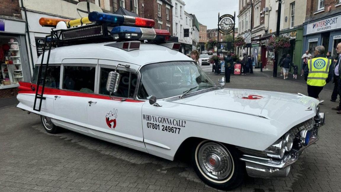 A white vintage American ambulance, decorated for its role in Ghostbusters, is parked in the square in Wellington with the town clock behind it