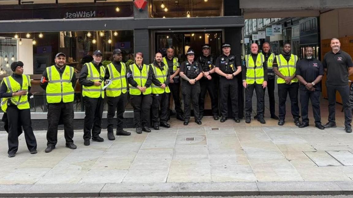 A picture of some of the security guards, there are more than 10 of them standing outside a coffee shop in Bedford, and there are three policeman with them. Most of the security guards are wearing high vis jackets.