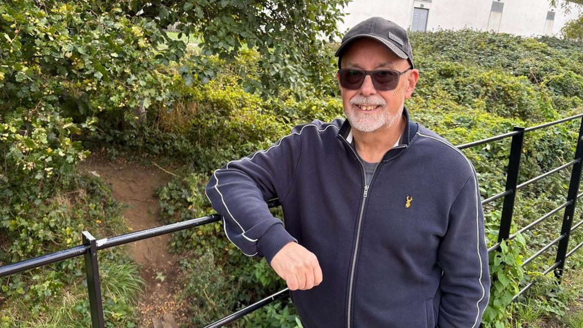 Nick DuGard with a short white beard wearing a grey cap and black zip-up top leading against a black metal railing. On the other side of the railing is a small embankment with a dirt path passing through foliage.