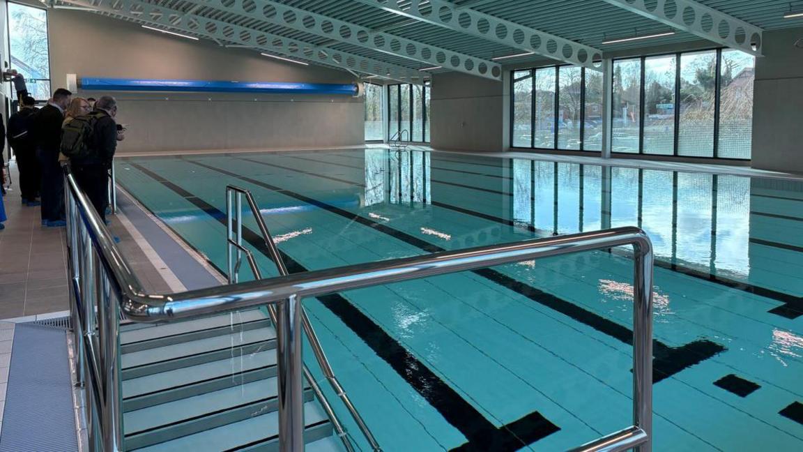 A swimming pool viewed from a balcony with metal rails around it and windows on the far side.