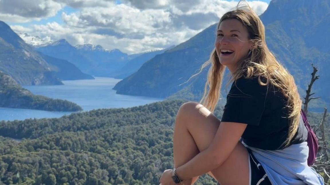 Charlotte on a hike. She is sitting smiling at the camera and in the background are mountains and a lake, with thick forest in the foreground