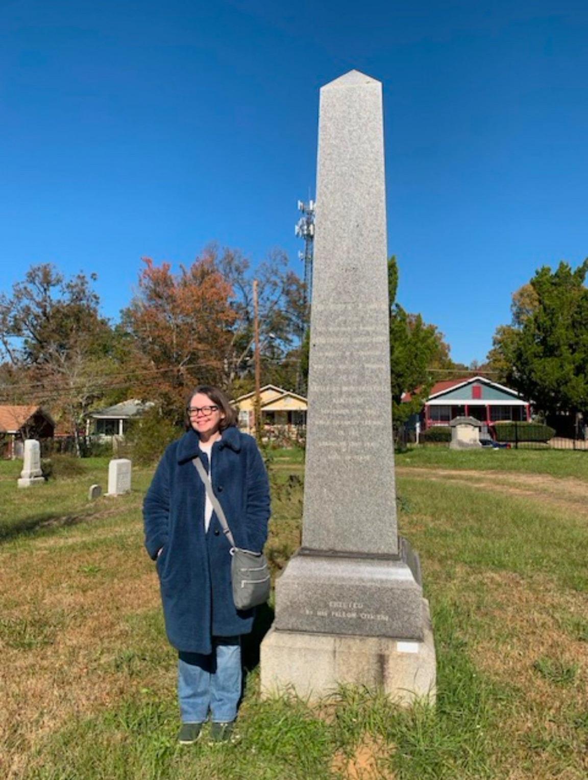 Lydia Melton pictured next to a tall grey obelisk. The monument is surrounded by grass and houses are visible in the background. Lydia has dark long hair and is wearing glasses. She is wearing a long, warm coat and has a bag over her shoulder. 