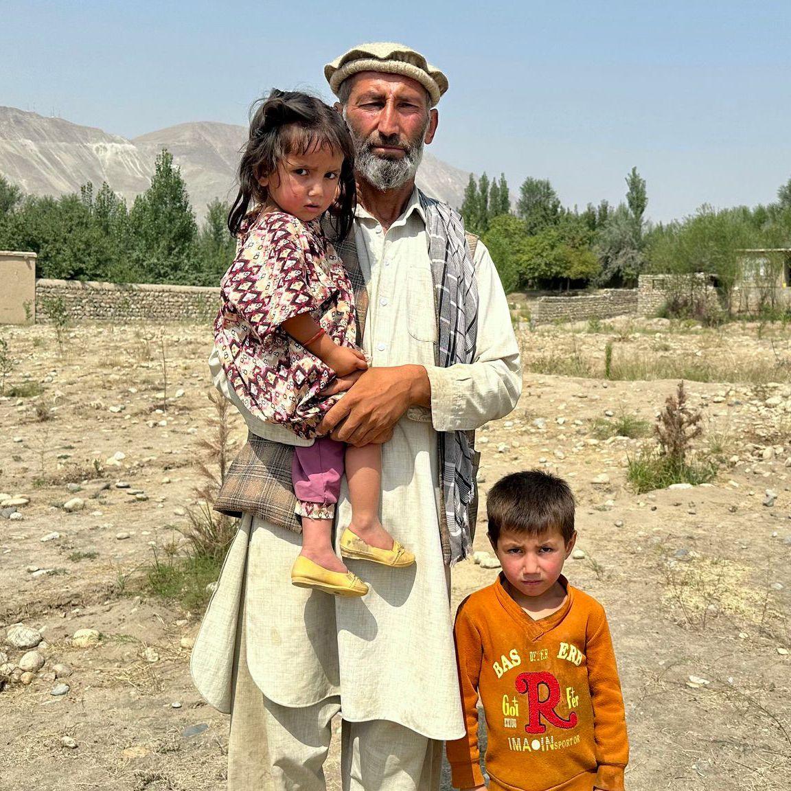 Abdul Wakeel stands looking at the camera while holding his daughter and with his young son standing next to them, against a rural backdrop and blue sky in Shesh Pol in the north-eastern Badakhshan province of Afghanistan.