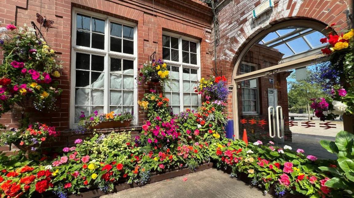 Red, pink, yellow and purple flowers in flower beds and in hanging baskets surround windows and an arched doorway at Bridlington station