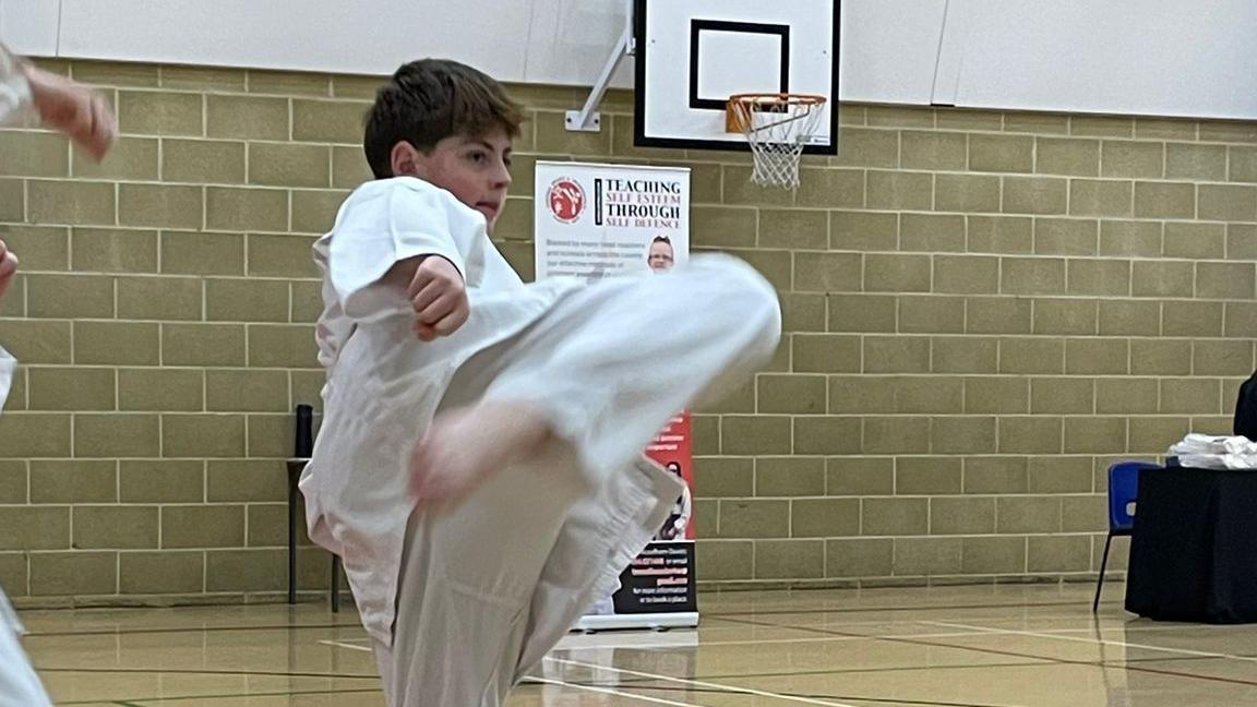 Archie during a training in a gym. He is photographed mid-kick. There is a basketball basket on the wall behind him.