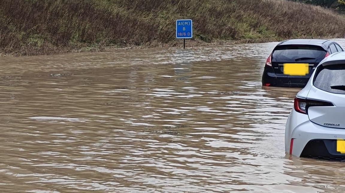 A blue sign reading A1(M) submerged in water. Two cars nearby are also submerged up to their number plates. 