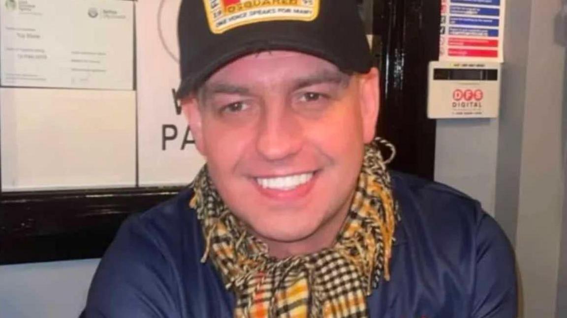 John Hardy is smiling into the camera. He's wearing a black cap, with a design on it, and a navy top and a checked scarf.