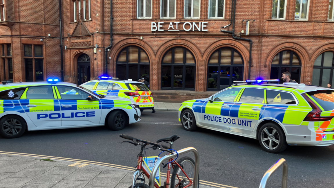 Three police cars are shown parked in the middle of a road to block traffic, with lights flashing on top of the vehicles. There is a nightclub in the background.