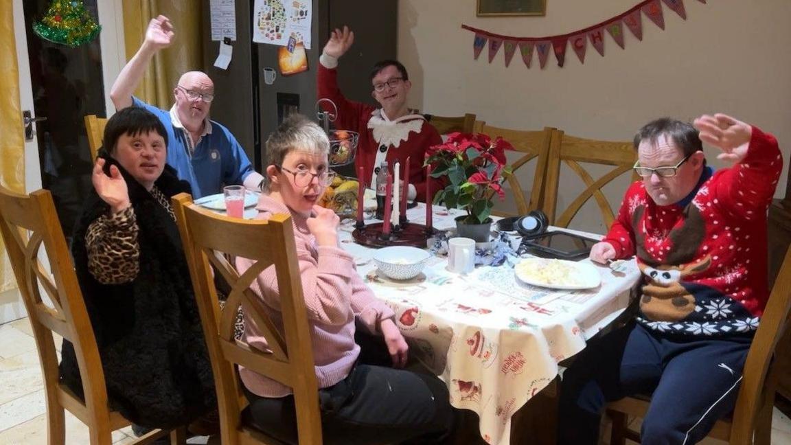 A family of five brothers and sisters are sitting around a dining room table. They are all looking towards the camera and waving. There are some Christmas decorations and candles on the table.