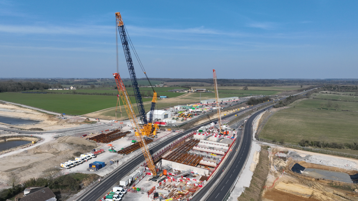 An aerial view of a construction site with cranes over a long stretch of dual carriageways. 