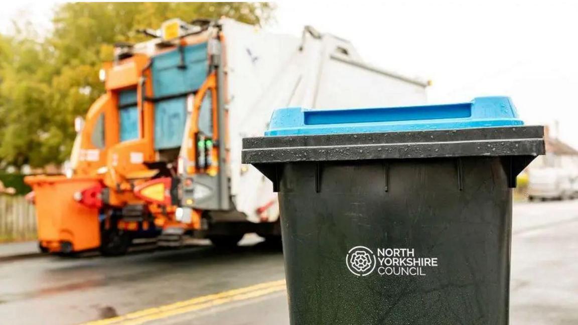 A wheelie bin with North Yorkshire Council logo on the side. A bin collection lorry in the background.