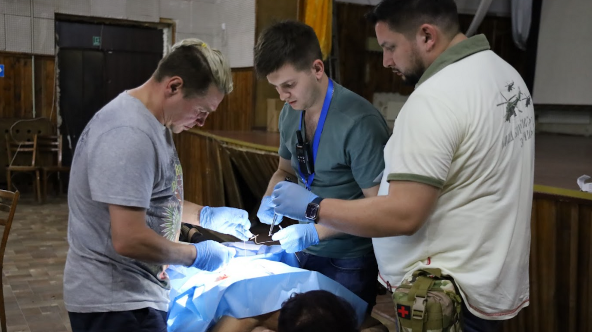 A woman lies face down on a table with a blue operating sheet over her body with three medics wearing gloves standing around her.