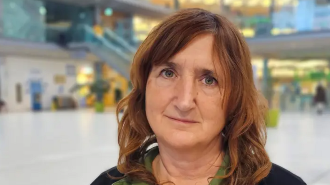 Lucy Galvin looks directly at the camera, unsmiling. Se has brown hair and is wearing a green top with dark cardigan. She has shoulder-length brown hair. She is standing in a glass atrium.