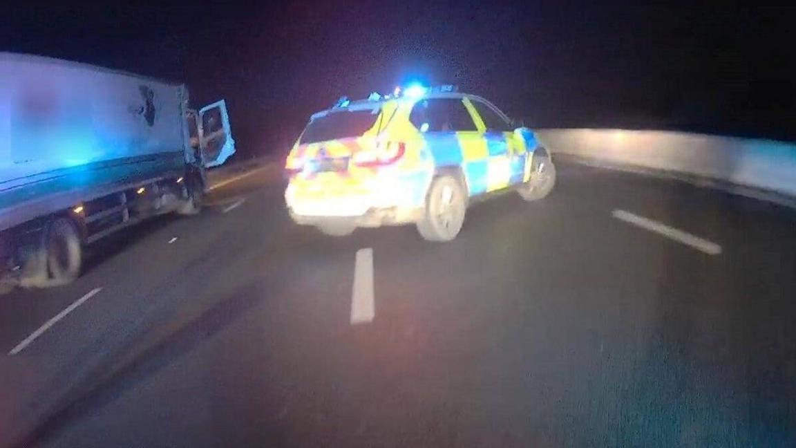 A police video still of a lorry parked in a lane of the M23, with a police patrol car parked beside it. The lorry cab's door is open.